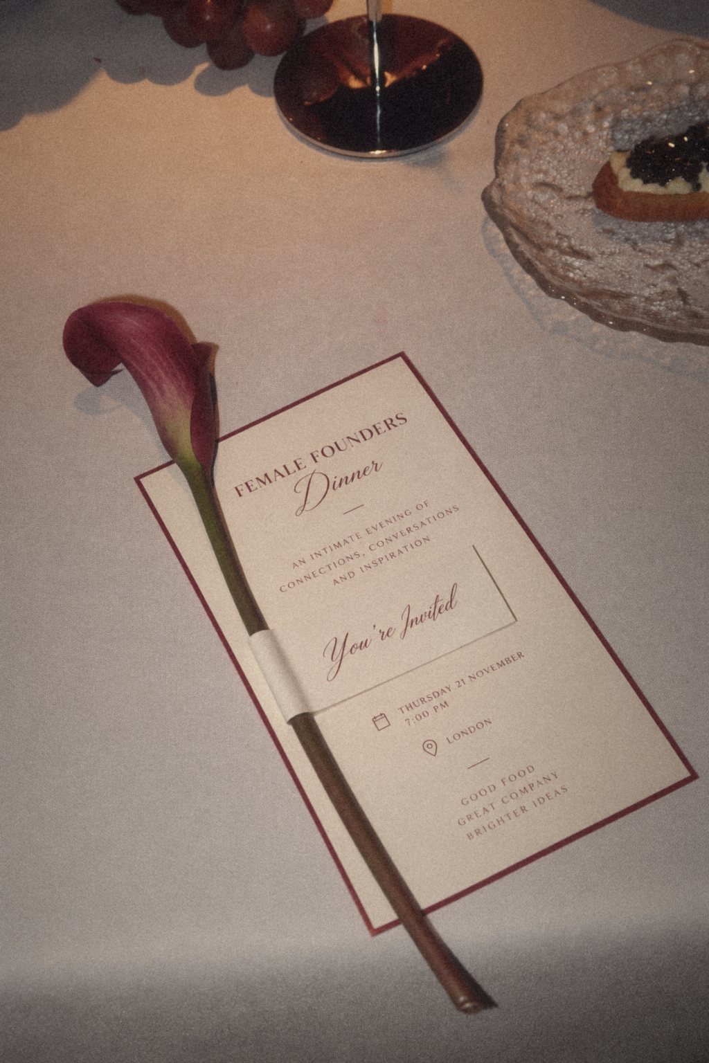 A cream Female Founders Dinner invitation card with a burgundy border and a single calla lily laid across it
