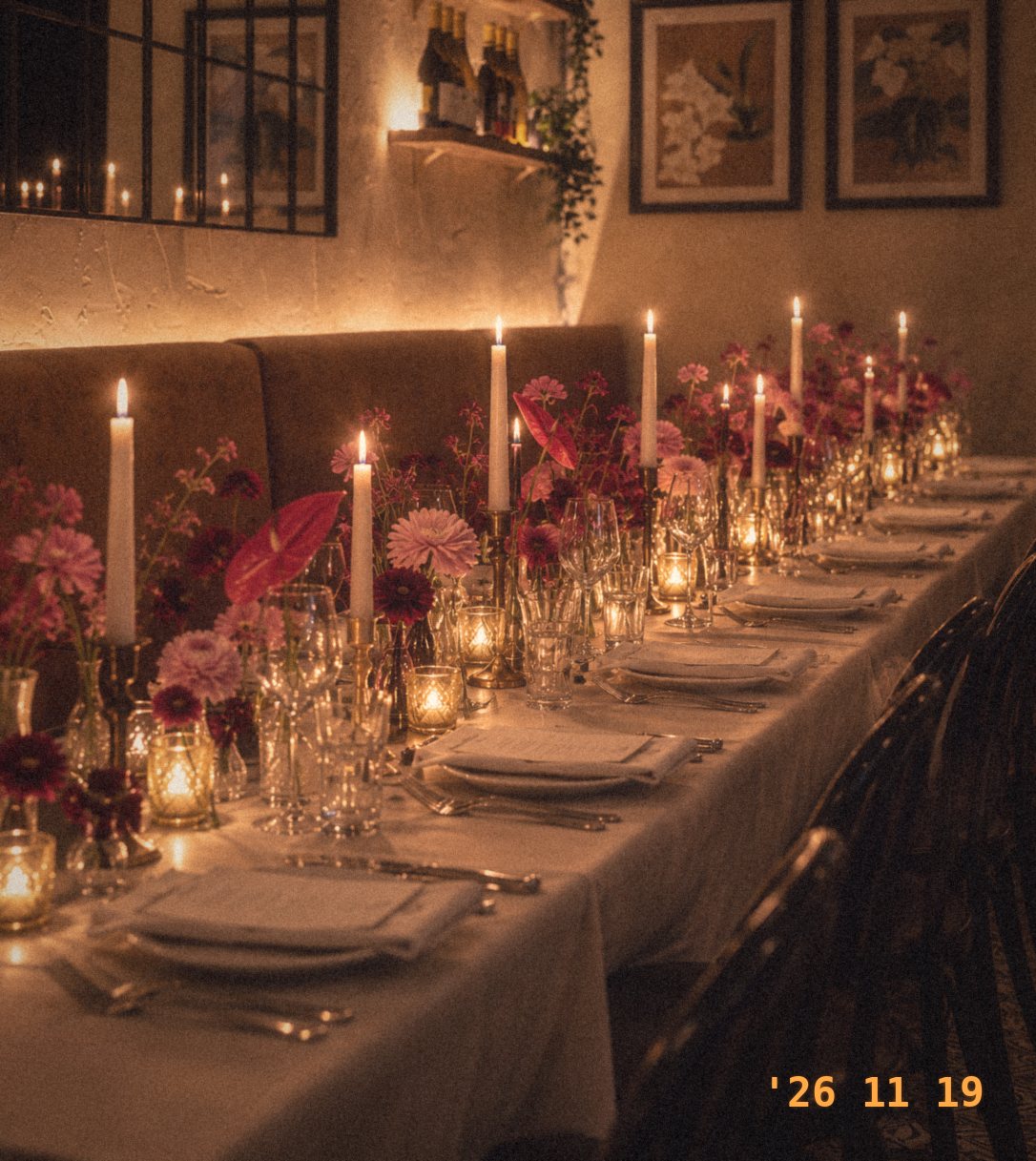 A long candlelit table set with white candles, deep pink and burgundy flowers and glassware, beside a velvet banquette